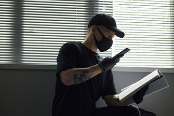 Young man wearing black mask and gloves sitting by window in company office holding smartphone in one hand and folder with confidential business document, scanning or photographing pages