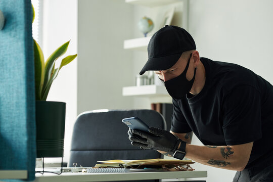 Young man wearing black cap and face mask leaning over desk using smartphone to take photos of confidential documents and business contacts in diary in office, black gloves on hands