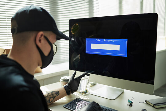 Young Caucasian man wearing black face mask and gloves sitting at desk using desktop computer, entering password on login screen to assess corporate network and steal confidential files