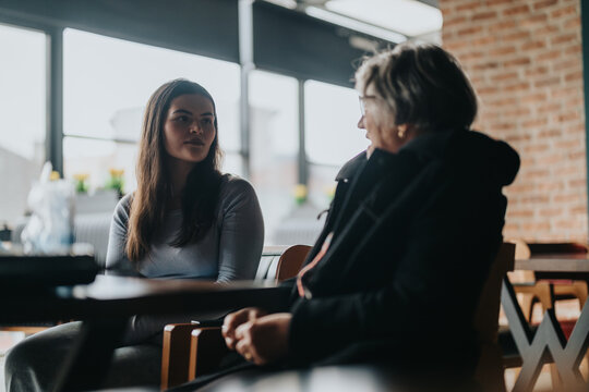 Two women, one young and one senior, having a thoughtful conversation in a warmly lit cafe with brick walls and large windows, creating a serene and engaging atmosphere.