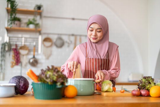 Muslim woman happy cooking healthy meal in kitchen wearing hijab making soup in pot with fresh vegetables food preparation activity nutrition, ramadan healthy recipe preparation, islamic food culture