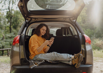 Happy young woman using smartphone sitting in open trunk of car during travel
