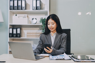 Young, smiling Asian businesswoman holding a jacket on her shoulder while using a mobile phone,...