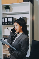 Young, smiling Asian businesswoman holding a jacket on her shoulder while using a mobile phone,...