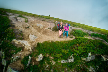Portrait of two hikers, equipped with backpacks and trekking poles, stand on rocky, green hillside....