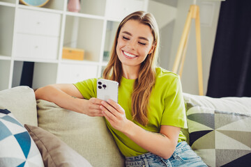 Pretty woman in casual attire smiling while using a smartphone and relaxing on a comfortable sofa indoors