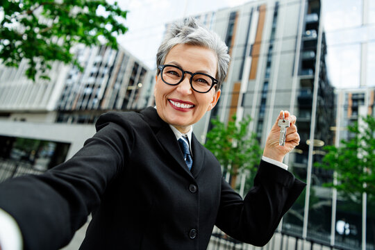 Cheerful professional businesswoman holding keys outdoors in city urban environment with modern buildings behind