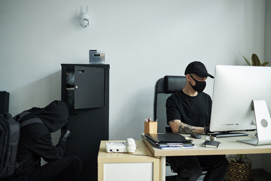 Caucasian male hacker in black cap and face mask breaking into office computer, while accomplice in hoodie raiding safe with confidential documents during burglary