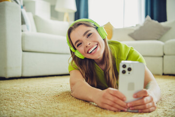 Cheerful woman in green heading wearing headphones, enjoying and smiling while holding smartphone...