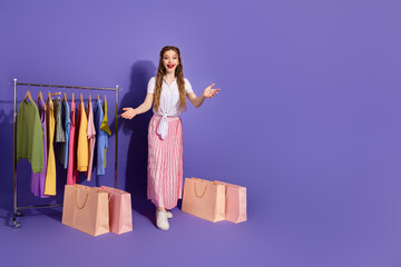 Young woman with shopping bags and clothing on a rack against a purple background, showcasing style...