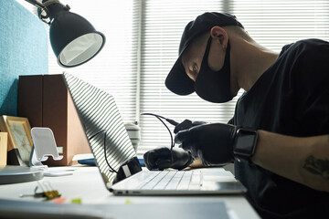 Male hacker wearing black cap and face mask sitting at desk in office, breaking into corporate...