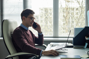 Caucasian young man sitting at desk talking on smartphone while working on laptop in modern office...