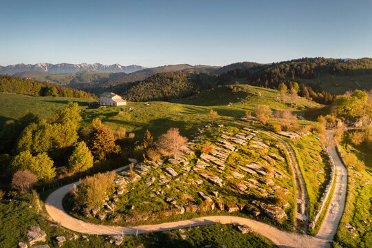 View of a winding road cutting through the rocky, green hills under a clear sky, with a lone building nestled in the distance, Parco Naturale Regionale della Lessinia, Veneto, Italy.
