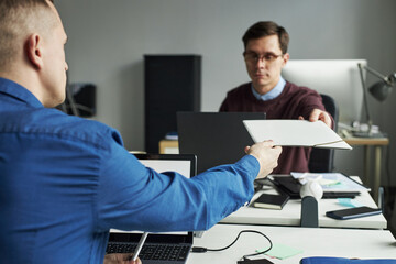 Fototapeta premium Caucasian man handing document folder to another male colleague sitting at desk in modern office, both focused on exchanging paperwork during business meeting. collaborating on presentation