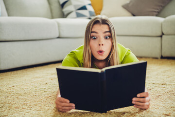 Young woman in casual attire lying in a cozy living room reading a captivating book while...