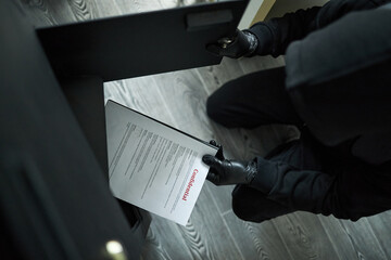 Unrecognizable male thief wearing black gloves and hoodie kneeling on floor, stealing confidential...