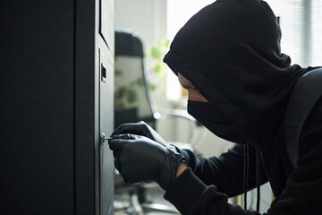 Young masked male intruder wearing black hoodie and gloves picking lock on large safe in office environment, face partially covered with mask, focused on breaking in to steal documents