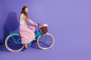 Young woman riding a vintage bike with a basket of flowers against a violet background in stylish...