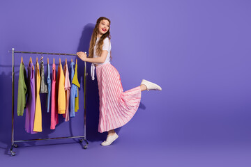 Young woman in stylish outfit standing by clothing rack against a vibrant purple background in a studio setting