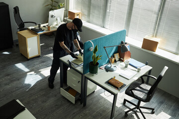 Young Caucasian male burglar standing at modern office desk searching through work documents,...