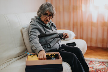 An elderly woman wearing glasses and a hoodie arranges belongings in a box. She sits on a cozy...