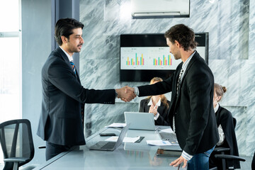 Business professionals shaking hands across conference table to finalize corporate agreement, multiracial team members applauding in background, office meeting showing success and teamwork