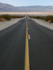 Fototapeta premium A long, straight road cuts through a vast desert valley under a clear blue sky, creating a striking sense of depth and endless perspective. Ideal for travel and adventure themes.