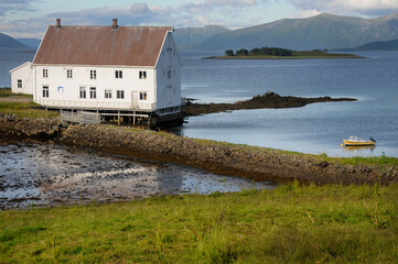 A white wooden house stands by a calm fjord in Norway, with a small yellow boat floating in front,...