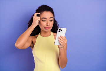 Young stylish woman in a yellow dress holds a smartphone with a thoughtful expression against a...