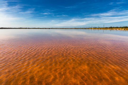 View of a vibrant pink salt lake reflecting the clear blue sky and wispy clouds, creating a surreal and colorful landscape, Saline di Cervia, Emilia-Romagna, Italy.