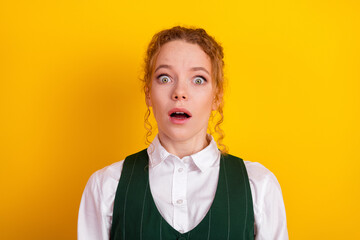Shocked young student with curly red hair in a uniform against vivid yellow background conveying...