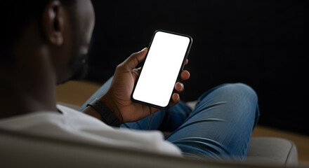 African american man holding smart phone with mockup white blank display, empty screen for app ads sitting on couch at home. Mobile applications technology concept, over shoulder close up view.