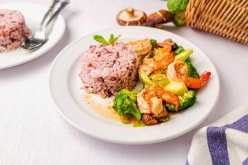 Stir fried broccoli with shrimp,shitake mushroom and riceberry in a white ceramic plate witn spoon and fork on white table background, clean food concept