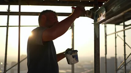 Skilled Worker Painting High on Scaffolding Against a Golden Hour Sunset, Industrial Construction Site Silhouette