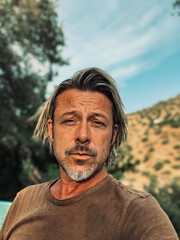 Tanned man with blond half long hair and grey stubble beard in brown t-shirt enjoys sunny summer...