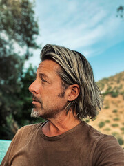 Tanned man with blond half long hair and grey stubble beard in brown t-shirt enjoys sunny summer...