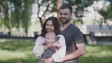 A joyful family stands together in a park, with the mother holding their baby and the father smiling beside them, all basking in the warm sunshine.