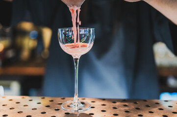 Bartender pours vibrant pink cocktail through a strainer into a glass at a stylish bar, showcasing professional mixology and drink presentation.