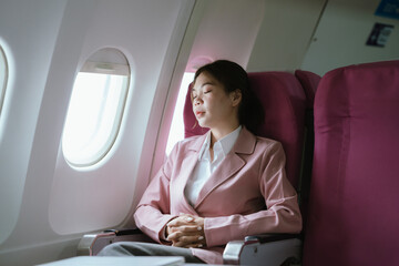A woman in a pink blazer sits by the airplane window with eyes closed, relaxing peacefully during...