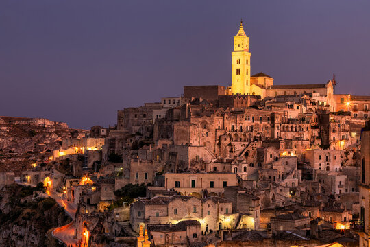 View of ancient stone buildings cascade down the hillside, bathed in warm light beneath a twilight sky, dominated by a glowing church tower, Sassi di Matera, Basilicata, Italy.