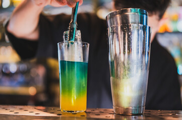 Close-up of bartender preparing colorful layered cocktail with ice using tongs. Glass, shaker, and...