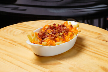 Fries with cheddar cheese and bacon in a white bowl on a wooden table.