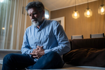 Mature man suffering from strong abdominal pain while sitting on sofa in living room during evening...