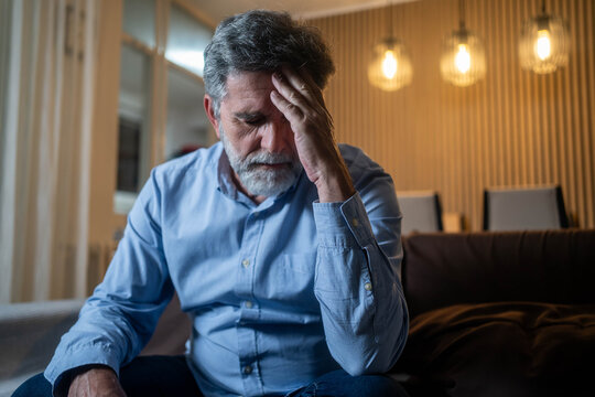 Mature man with headache holding forehead in pain, sitting in living room during evening with pained expression