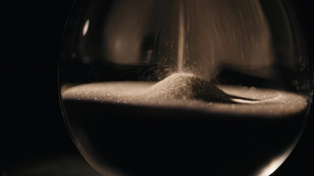 Closeup sand falling hourglass standing dark room. Gold grains flowing inside
