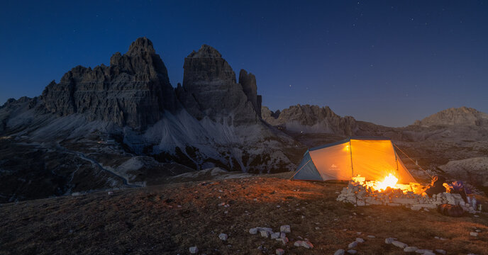 View of a glowing tent, nestled among jagged peaks under a starlit sky, with a warm campfire crackling invitingly in Tre Cime di Lavaredo, Trentino-Alto Adige, Italy.