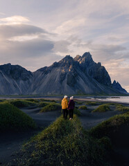 Fototapeta premium Two travelers explore lush grass and black sand dunes beneath Vestrahorn mountain in Stokksnes, Iceland, embodying adventure in a stunning twilight landscape.