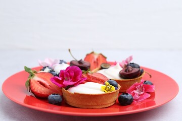 Sweet tartlets with berries and flowers on grey table against light background, closeup. Delicious dessert