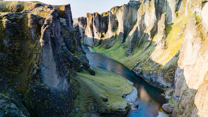 Majestic cliffs rise above the shimmering river in Fjadrargljufur Canyon in Iceland, showcasing Icelands dramatic natural beauty during golden hour. Lush green moss blankets rocky formations.