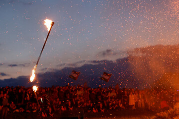 Fiery spectacle at night with two flaming torches held high above a large crowd, sparks raining...